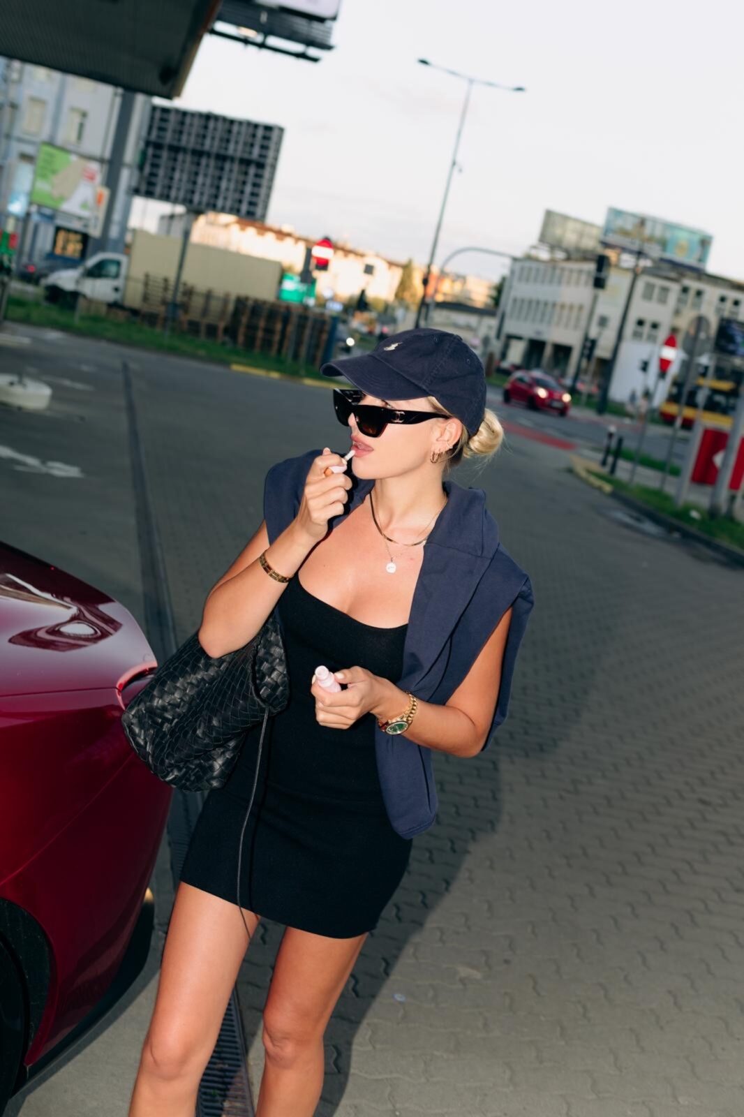 Izabella Budryn kim jest: Kobieta w sportowym, miejskim stylu: mała czarna sukienka na ramiączkach, czapka z daszkiem i ciemne okulary przeciwsłoneczne. Stylizacja podkreślona dużą, czarną torebką i złotymi dodatkami, idealna na casualowe wyjście.