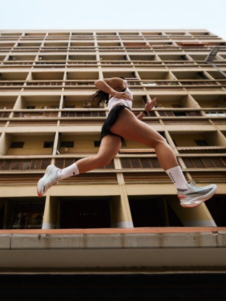 Puma Go Wild: Sportowa sylwetka kobiety uchwycona w dynamicznym skoku podczas biegu. Zdjęcie wykonane od dołu, z perspektywy podkreślającej nowoczesny budynek w tle, tworząc efektowną kompozycję podkreślającą szybkość i lekkość ruchu.