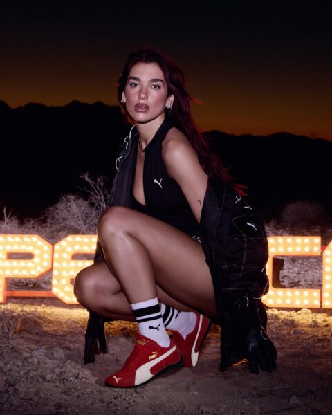 Dua Lipa pozująca na tel wielkiego neonu w butach PUMA Speedcat 2025. Piosenkarka jest ubrana w body i rajstopy oraz płaszcz i kuca na ziemii