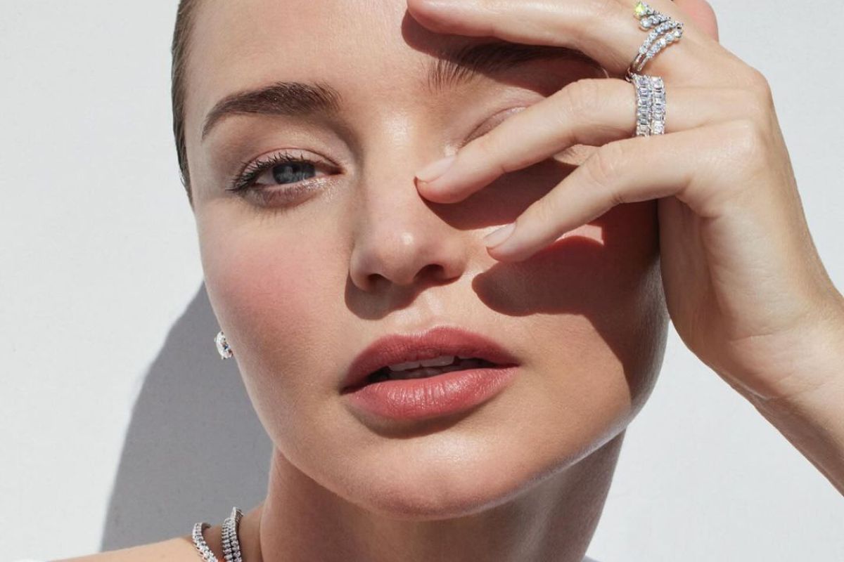 Modelka Miranda Kerr zasłaniająca twarz dłonią ozdobioną pierścionkami. Ma na sobie biały top i kilka naszyjników.