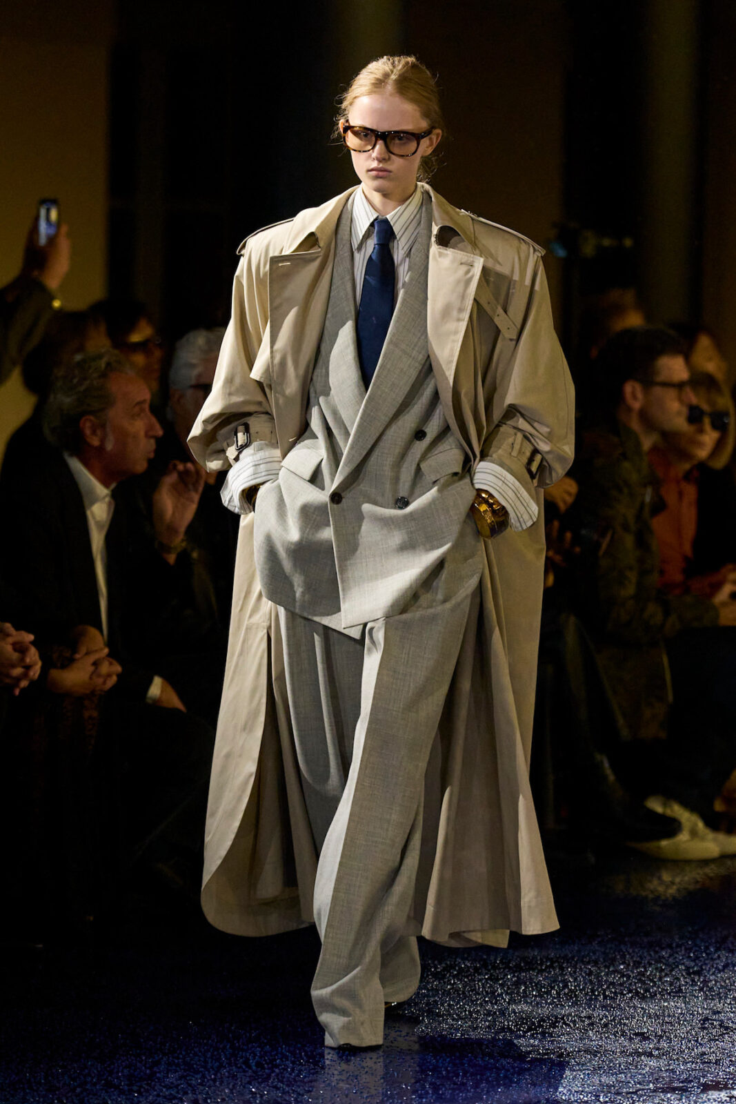 Zdjęcie przedstawia modelkę na wybiegu, ubraną w stylizację o klasycznym, eleganckim charakterze. Ma na sobie dwurzędowy, oversize'owy szary garnitur z szerokimi spodniami oraz kremowy trencz narzucony na ramiona. Stylizację dopełniają duże okulary o ciemnych oprawkach, granatowy krawat i złota biżuteria, tworząc nowoczesną interpretację formalnej elegancji.