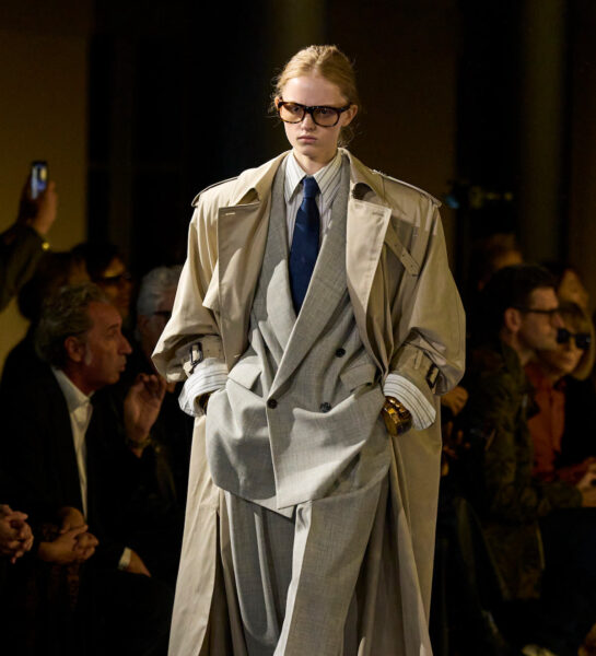 Zdjęcie przedstawia modelkę na wybiegu, ubraną w stylizację o klasycznym, eleganckim charakterze. Ma na sobie dwurzędowy, oversize'owy szary garnitur z szerokimi spodniami oraz kremowy trencz narzucony na ramiona. Stylizację dopełniają duże okulary o ciemnych oprawkach, granatowy krawat i złota biżuteria, tworząc nowoczesną interpretację formalnej elegancji.