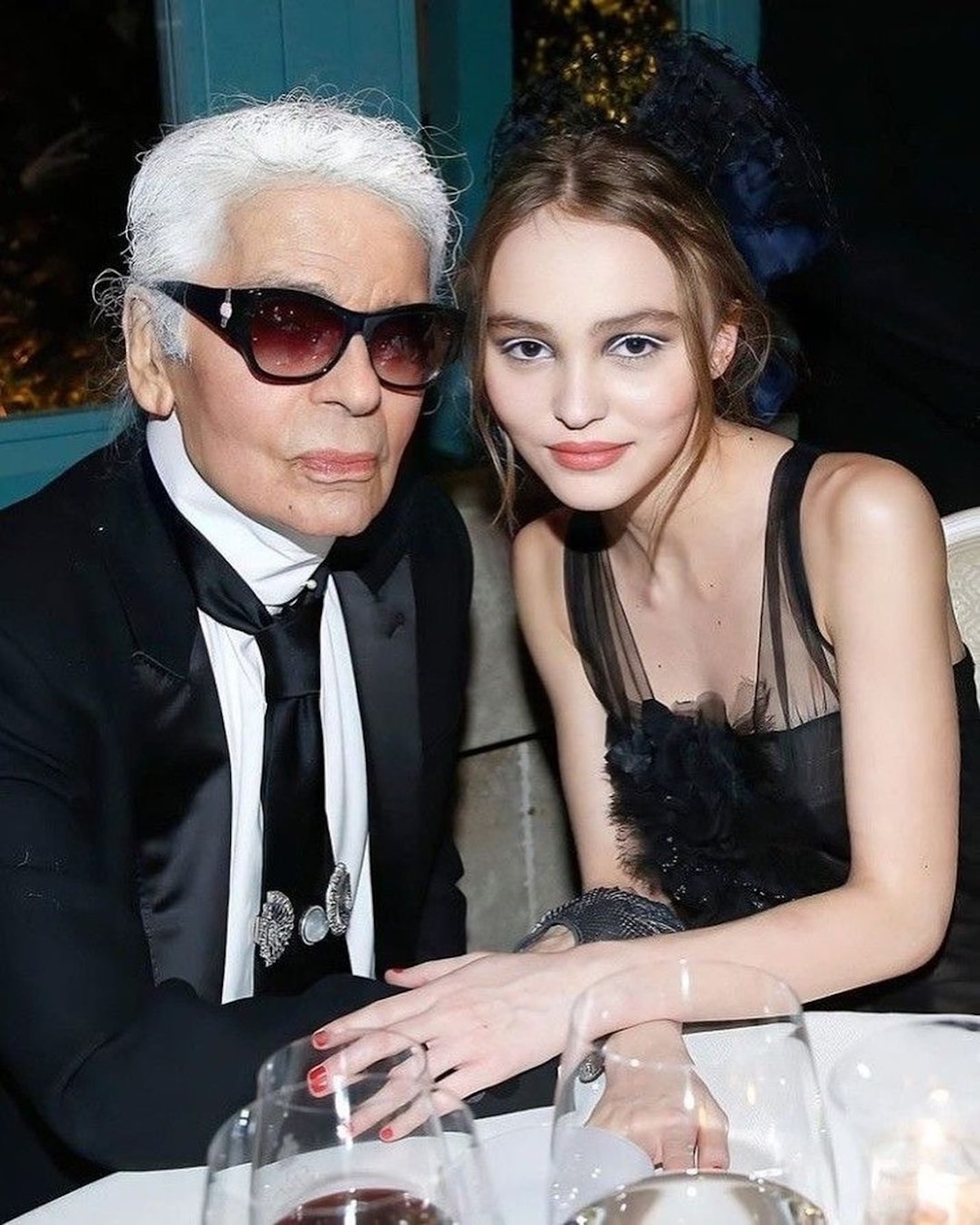 Projektanci i ich znane muzy: Lily Rose-Depp i Karl Lagerfeld na zdjęciu. Trzymają się za ręce siedząc przy jednym stoliku, widać, że łączy ich niezwykła więź