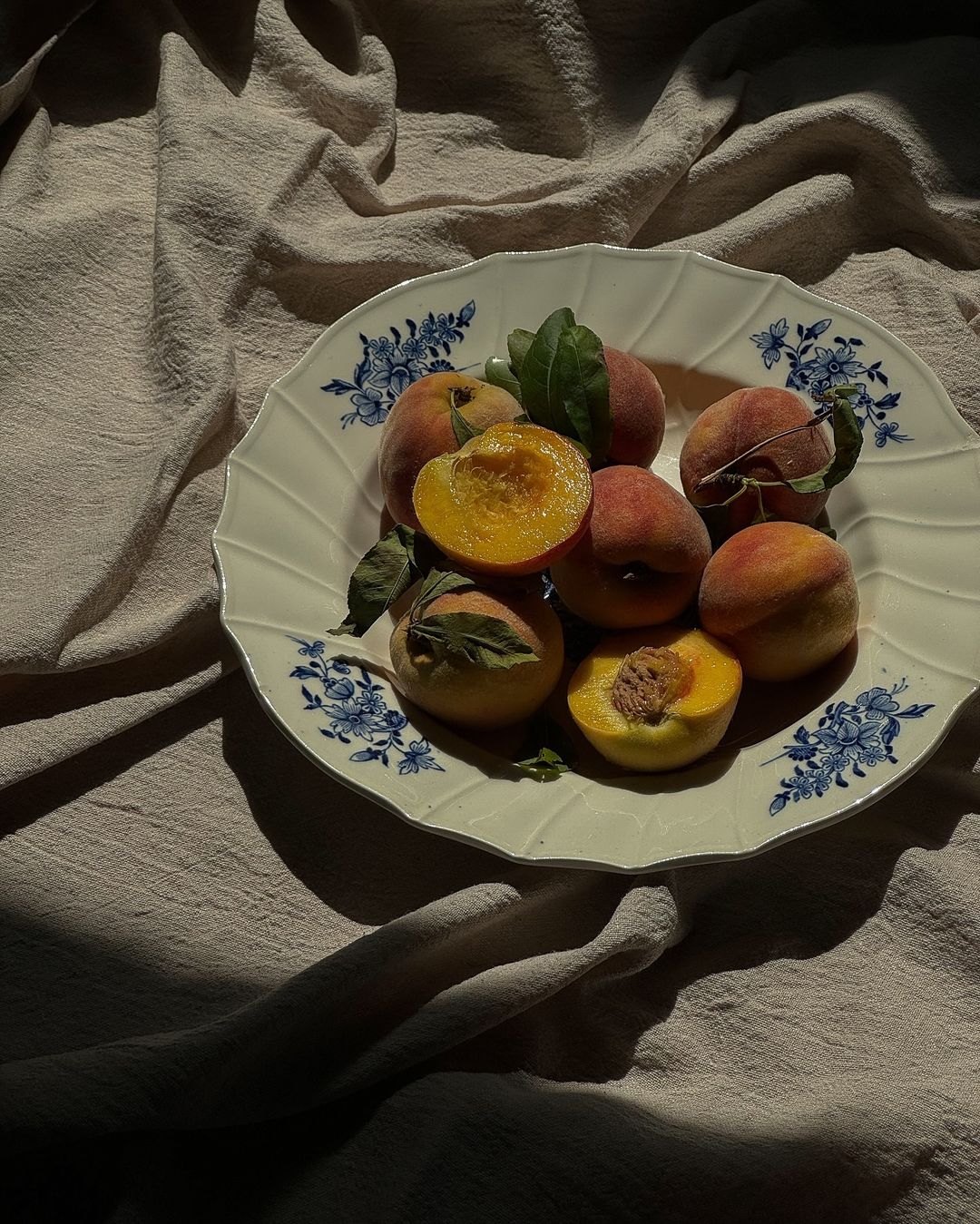Dieta na piękną skórę to przede wszystkim warzywa. Na zdjęciu, na talerzu leżą brzoskwinie rozkrojone w pół. Talerz ma niebieskie zdobienia i postawiony jest na nierówno ułożonym obrusie. Na miskę z owocami pada struga słońca