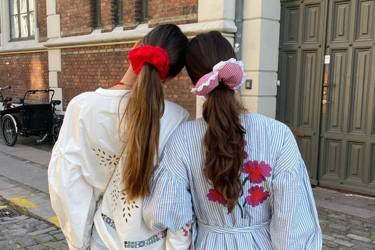 Dwie kobiety z długimi włosami związanymi w niski kucyk kolorowymi gumkami typu scrunchie. Mają na sobie koszule, stoją tyłem do obiektywu. Znajdują się na tle starego budynku,