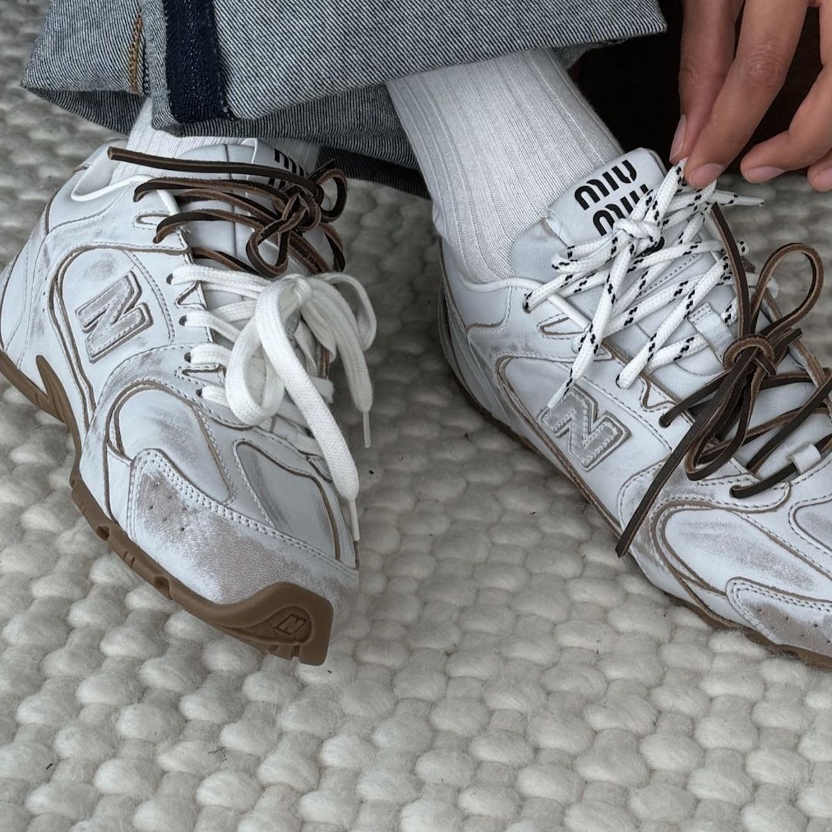 Zdjęcie przedstawia stylowe sneakersy, będące efektem współpracy Miu Miu i New Balance, wyróżniające się minimalistycznym designem z białą bazą i brązowymi akcentami. Na języku buta widoczne jest logo "Miu Miu", podkreślające ekskluzywny charakter modelu. Buty są ozdobione różnymi sznurowadłami – jedno białe, a drugie brązowe – co nadaje im modny, niekonwencjonalny wygląd. W tle widać fragment dżinsów oraz jasne skarpety, które dopełniają casualowo-elegancki styl.