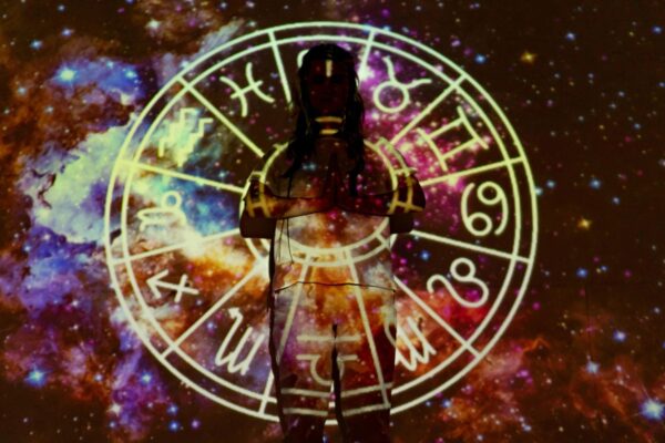 Koło reprezentujące znaki zodiaki i daty. Na ich tle stoi kobieta i trzyma złożone dłonie.