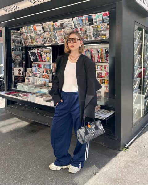 Młoda kobieta ubrana w nowoczesny streetwearowy styl pozuje przed kioskiem z gazetami. Ma na sobie oversizową czarną marynarkę, biały top, granatowe spodnie dresowe z trzema białymi paskami po bokach (marka Adidas) i masywne białe sneakersy. Dodatki to duże, prostokątne okulary przeciwsłoneczne, biżuteria w postaci łańcuszków oraz mała torebka z wzorem monogramu. Kiosk w tle wypełniony jest różnorodnymi magazynami, co podkreśla miejski klimat zdjęcia.