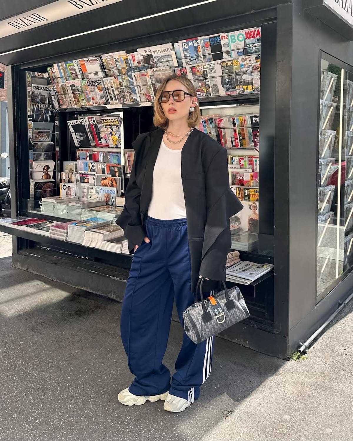 Młoda kobieta ubrana w nowoczesny streetwearowy styl pozuje przed kioskiem z gazetami. Ma na sobie oversizową czarną marynarkę, biały top, granatowe spodnie dresowe z trzema białymi paskami po bokach (marka Adidas) i masywne białe sneakersy. Dodatki to duże, prostokątne okulary przeciwsłoneczne, biżuteria w postaci łańcuszków oraz mała torebka z wzorem monogramu. Kiosk w tle wypełniony jest różnorodnymi magazynami, co podkreśla miejski klimat zdjęcia.