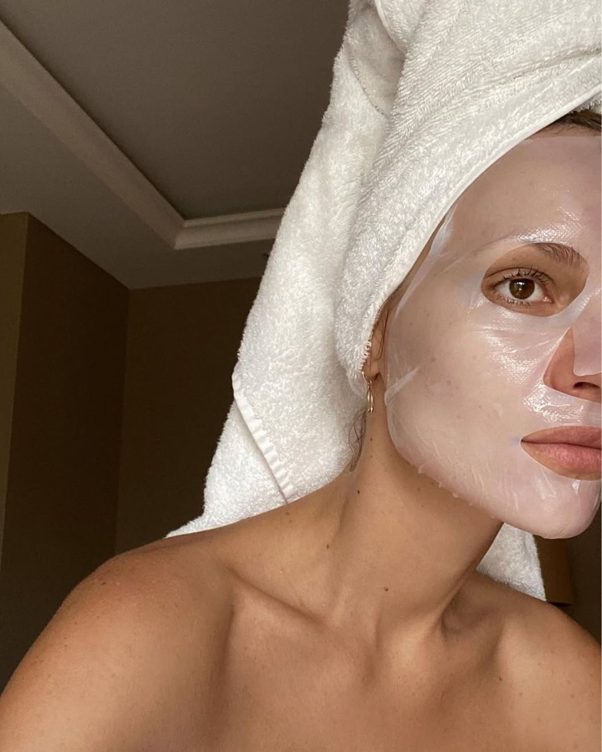 Kobieta z maseczką na twarzy i turbanem z białego ręcznika na głowie. Siedzi w pokoju hotelowym.