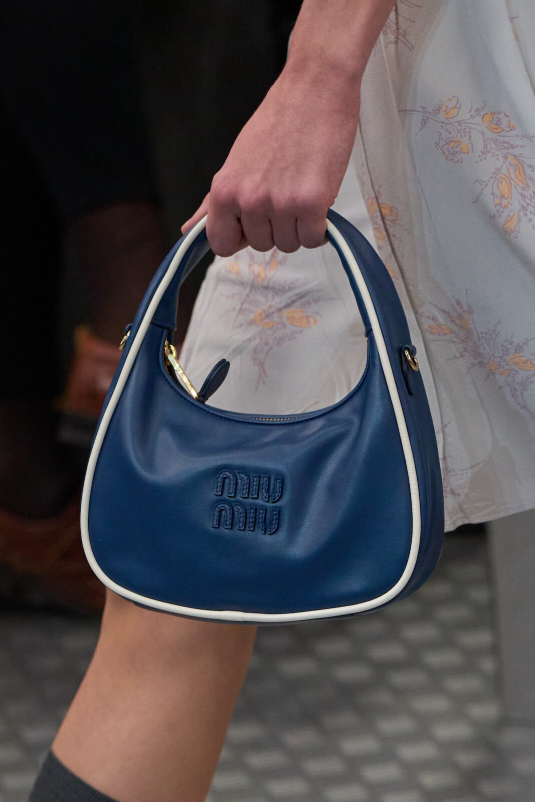 Osoba trzymająca niebieską torebkę Miu Miu typu hobo, z białymi krawędziami i tłoczonym logo marki z przodu. Torebka wykonana z gładkiej skóry, ma zaokrąglony kształt i krótką rączkę. W tle widoczny fragment zwiewnej sukienki z delikatnym, kwiatowym wzorem.