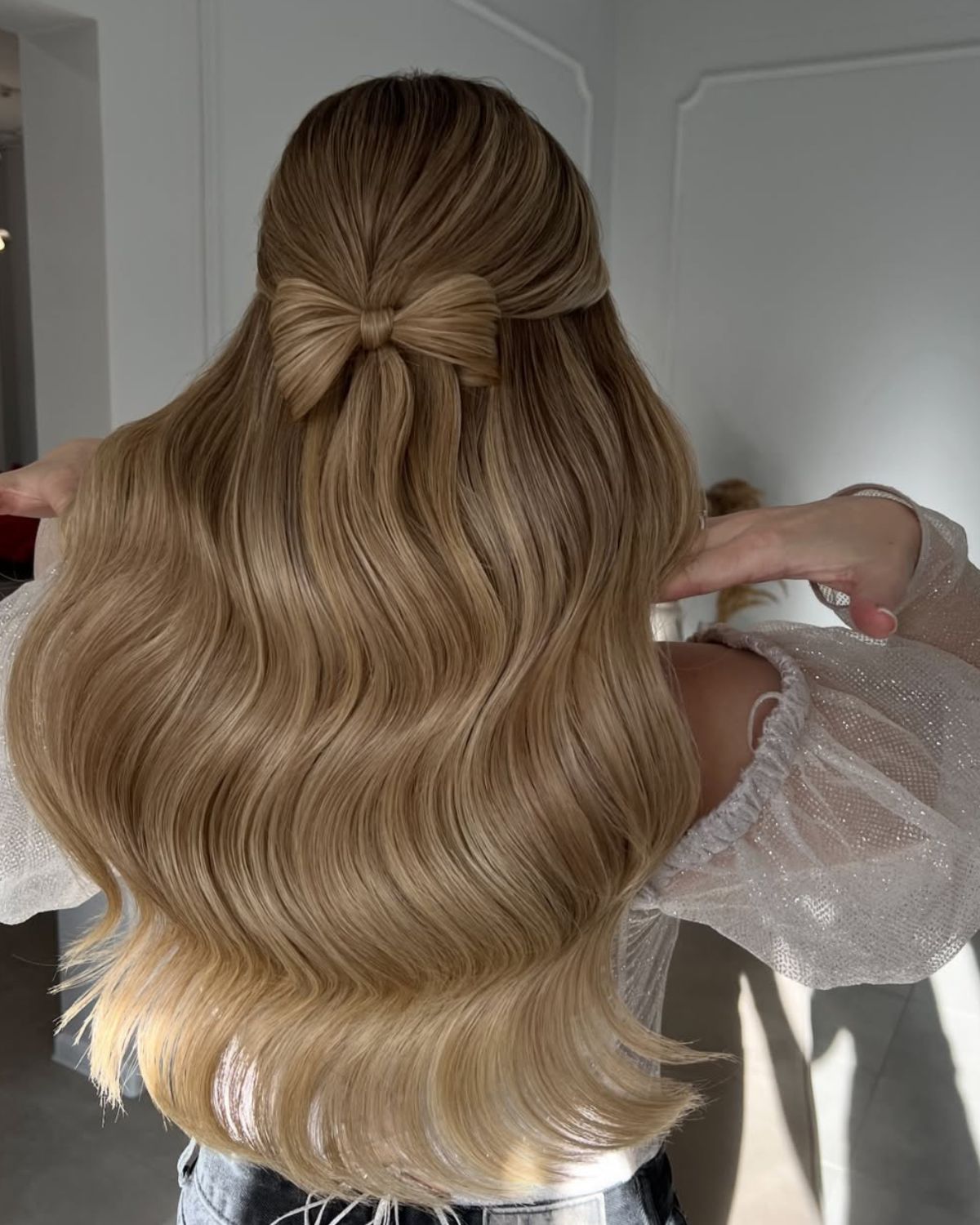 Kobieta stojąca tyłem prezentująca najmodniejsze odcienie blondu 2025, czyli miodowy blond. Włosy są gładkie, lśniące, sięgające poniżej łopatek. Kobieta ma półupięcie w kształcie kokardki. Ubrana jest w bluzkę z siateczki z brokatowym wykończeniem oraz spodnie jeansowe.