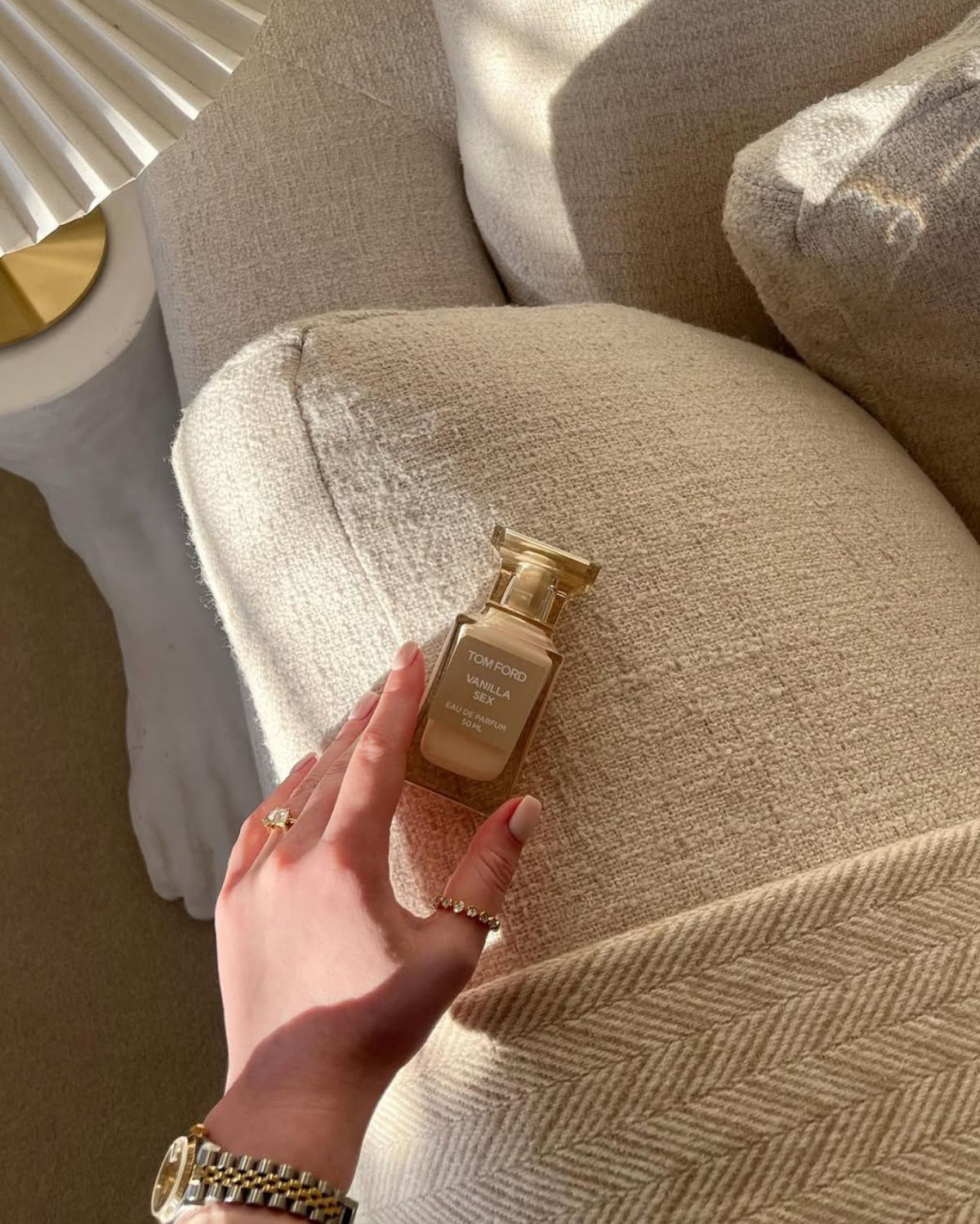 Perfumy marki Tom Ford jako najpiękniejsze perfumy na święta 2024. Chwyta je kobieca dłoń z zegarkiem i pierścionkami na palcach. Buteleczka perfum znajduje się na oparciu beżowej kanapy. Obok widać lampę stołową z karbowanym kloszem.