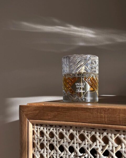 Perfumy w pięknym, kryształowym flakonie z tłoczonymi wzorami. SToją na rattanowej szafce w kolorze brązowym. Na perfumy pada cień, który tworzy efektowne refleksy na ścianie odbijające się od flakonu.