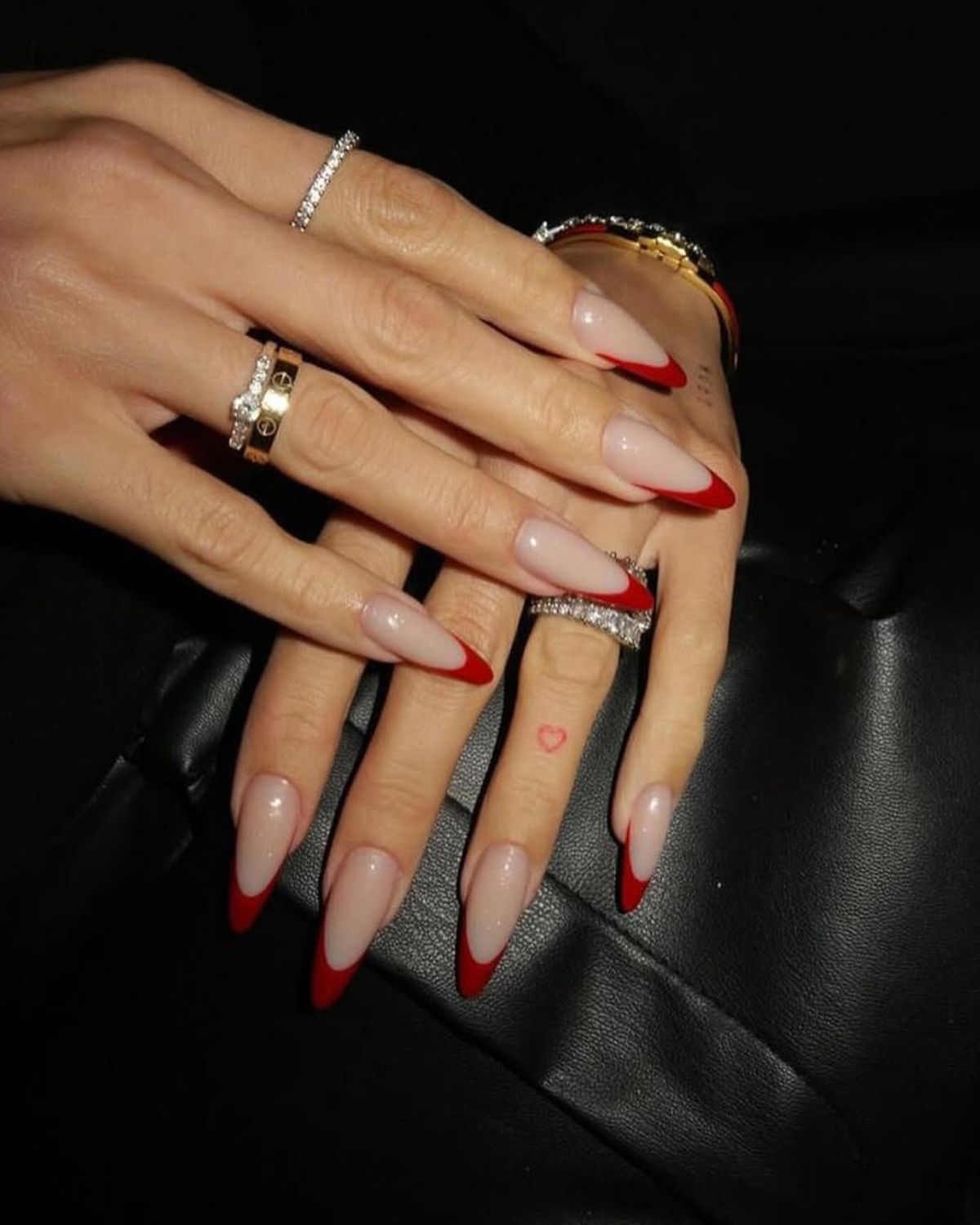 Dłonie z długimi paznokciami w kształcie migdała pomalowane w stylu french manicure z czerwoną końcówką. Na palcach znajdują się liczne pierścionki w kolorze złota wysadzane kamieniami. Dłonie układają się na czarnej torebce. W tle widać czarny płaszcz.