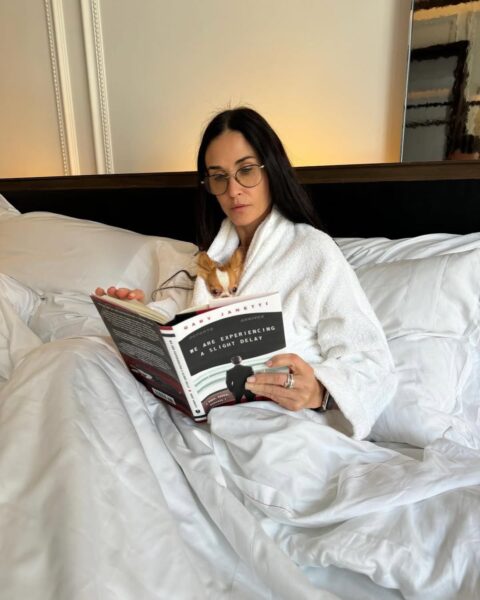 Na zdjęciu widać Demi Moore siedzącą na łóżku w białym szlafroku, z książką w rękach. Towarzyszy jej mały piesek rasy chihuahua, wtulony w jej ramiona. Książka, którą czyta, to "We Are Experiencing a Slight Delay" autorstwa Gary'ego Janettiego. Sceneria jest przytulna i wskazuje na spokojny moment relaksu.