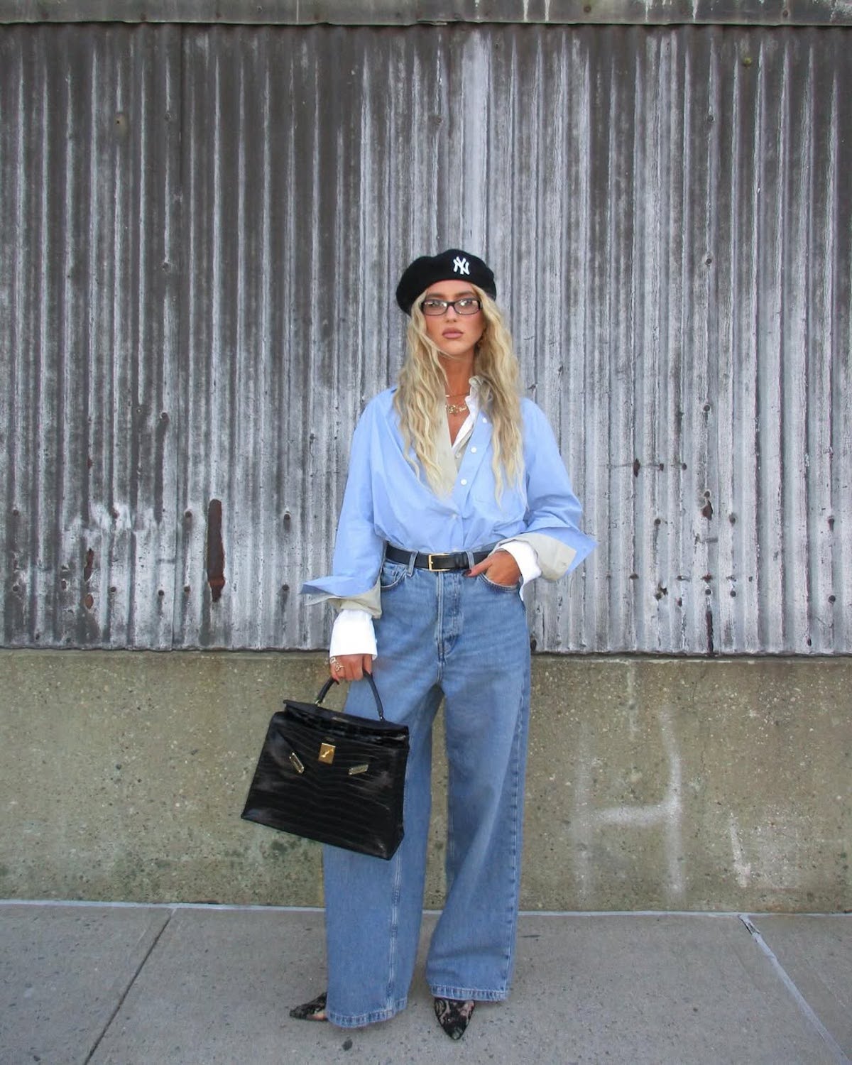 Stylizacja streetwearowa: modelka w niebieskiej koszuli oversize, modnych jeansach o szerokim kroju, czarnym berecie z logo NY i eleganckiej torebce, prezentuje casualowy szyk.