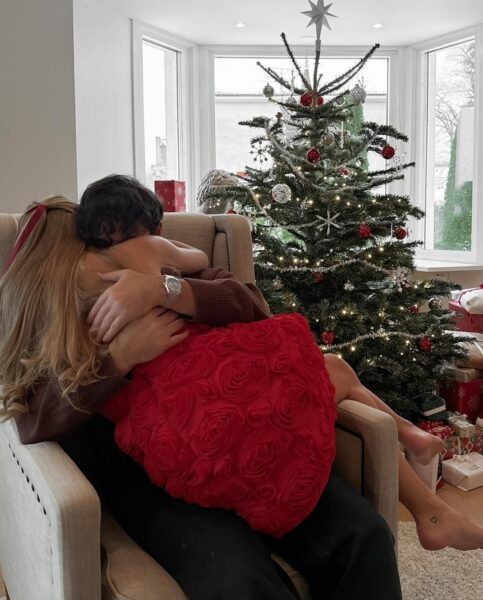 Para przytulająca się w świątecznym wnętrzu, w tle pięknie udekorowana choinka z ozdobami i prezentami. Kobieta w eleganckiej czerwonej sukni ozdobionej różanymi motywami, idealny pomysł na prezent dla kobiety na święta, podkreślający magię Bożego Narodzenia i ciepło domowej atmosfery.