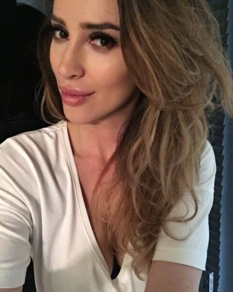 Ewelina Ślotała pogrzeb: kobieta w białym T-shircie, selfie z bliska, ukazujące jej perfekcyjny makijaż i fryzurę z długimi, falowanymi włosami.