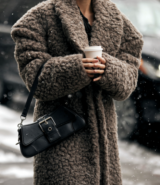 Elegancki zimowy look w stylu après-ski 2025 – kobieta w obszernej brązowej kurtce typu teddy coat, trzymająca kubek gorącego napoju. Stylizacja uzupełniona klasyczną czarną torebką na ramię z dużą klamrą. Tło z zaśnieżoną ulicą i zimowym krajobrazem podkreśla luksusowy klimat "old money".
