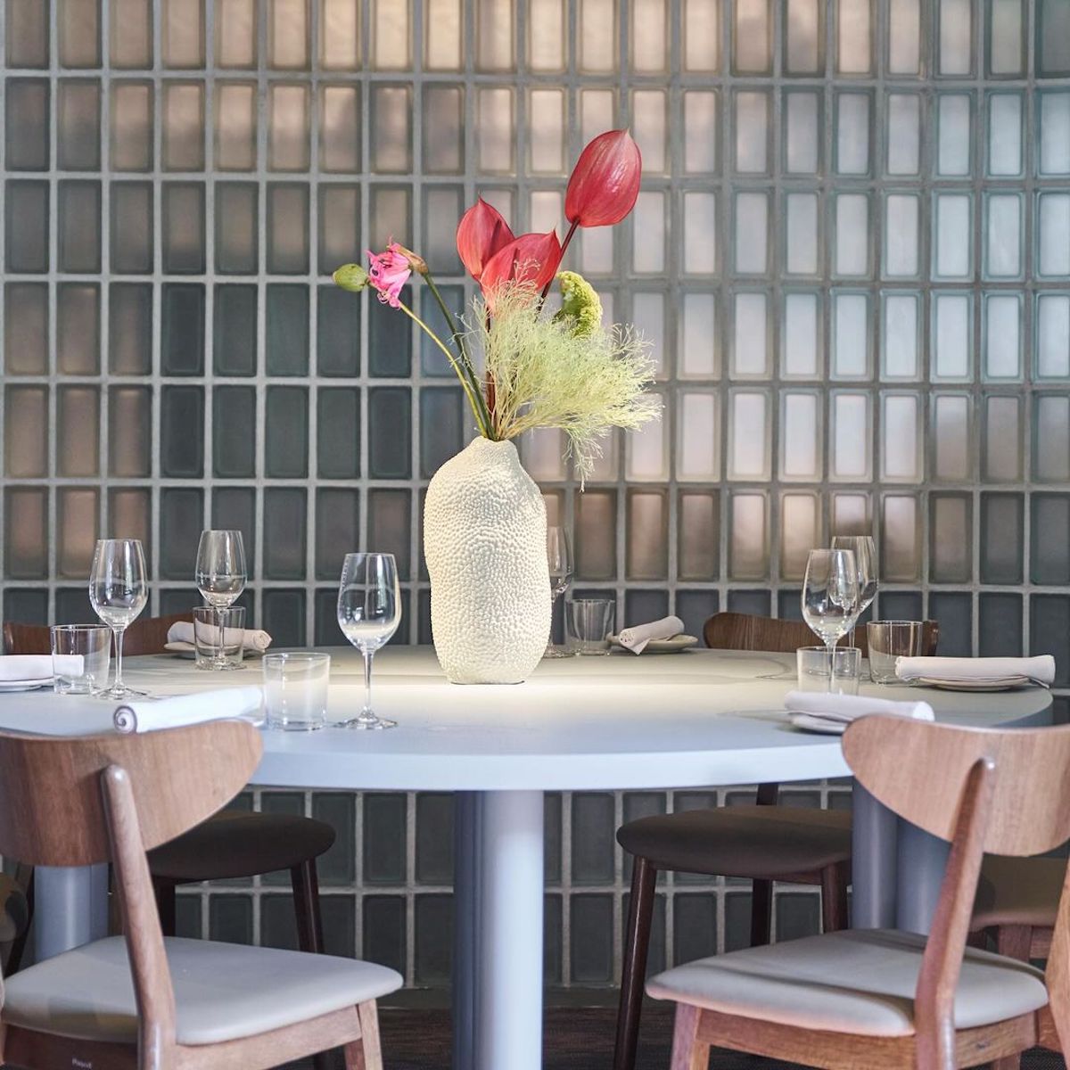 Zdjęcie przedstawia eleganckie wnętrze restauracji. Centralnym punktem jest okrągły stół o gładkiej, jasnej powierzchni, na którym znajduje się ozdobny wazon w nieregularnym kształcie, przypominający strukturę ceramiki, z kompozycją kwiatową. Kwiaty w wazonie to czerwone anturium.