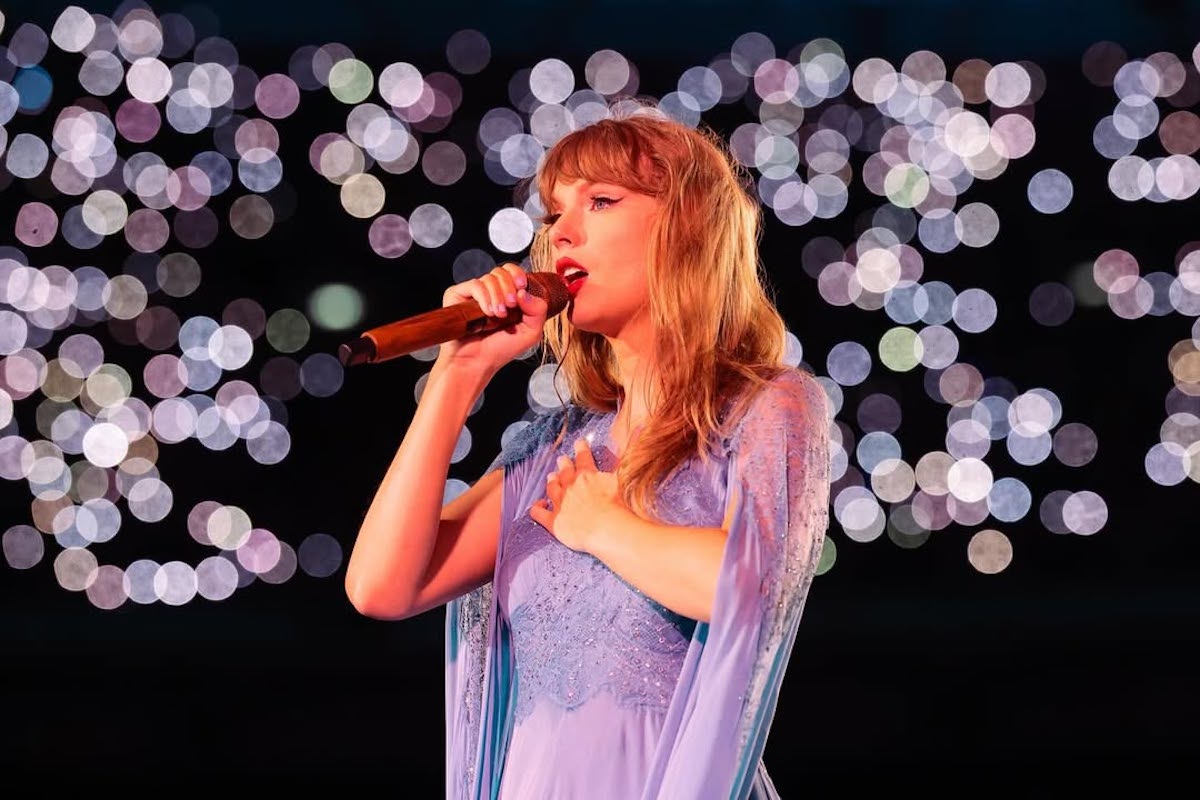 Widzimy Taylor Swift śpiewającą na scenie, trzymającą mikrofon w jednej ręce, a drugą dłoń przyłożoną do klatki piersiowej. Ma na sobie zwiewną, jasnofioletową suknię z delikatnymi zdobieniami i koronkowymi elementami. W tle widoczne są rozmazane światła, tworzące efekt bokeh, co nadaje obrazowi magicznej atmosfery. Scena wygląda na oświetloną, co podkreśla artystkę i jej emocjonalny wyraz twarzy.