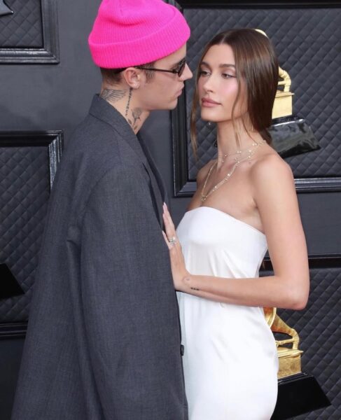 Para stoi razem na tle czarnej ściany z pikowanym wzorem, przed złotą statuetką Grammy. Kobieta ubrana jest w białą, elegancką, dopasowaną suknię bez ramiączek, a jej włosy są gładko zaczesane. Mężczyzna nosi różową czapkę beanie, ciemne okulary, szary oversize’owy garnitur i ma widoczne tatuaże na szyi. Patrzą sobie w oczy w bliskim ujęciu.