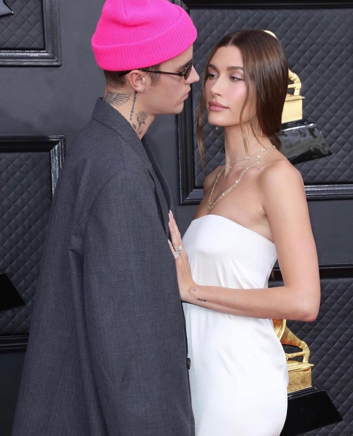 Para stoi razem na tle czarnej ściany z pikowanym wzorem, przed złotą statuetką Grammy. Kobieta ubrana jest w białą, elegancką, dopasowaną suknię bez ramiączek, a jej włosy są gładko zaczesane. Mężczyzna nosi różową czapkę beanie, ciemne okulary, szary oversize’owy garnitur i ma widoczne tatuaże na szyi. Patrzą sobie w oczy w bliskim ujęciu.