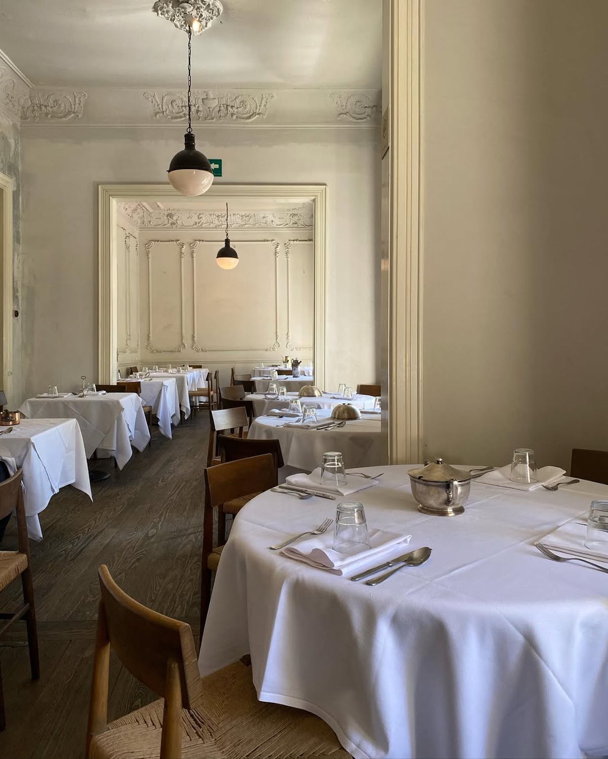 Elegancka restauracja w Warszawie, idealna na Walentynki, z białymi obrusami na okrągłych i prostokątnych stołach, ustawionych na drewnianej podłodze. Wnętrze ozdobione klasycznymi sztukateriami na ścianach i suficie, a nad stołami wiszą stylowe, czarne lampy. Jasne tonacje i minimalistyczny wystrój tworzą romantyczną atmosferę, doskonałą na wyjątkowy wieczór.