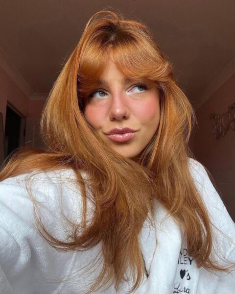 Kobieta w rudych, długich włosach rozpuszczonych na ramiona. Ma francuską grzywkę wywiniętą na zewnątrz w stylu wispy. Ubrana jest w białą, polarową bluzę.