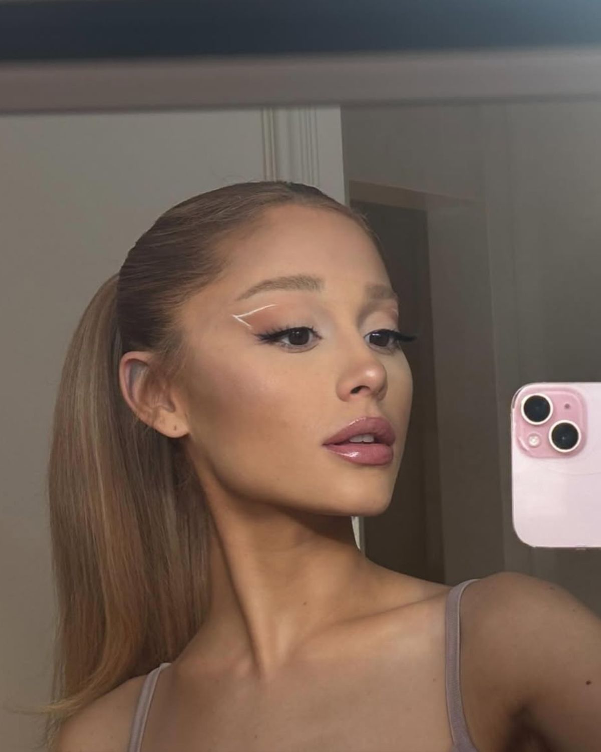 Aktorka i piosenkarka Ariana Grande prezentująca fryzurę z kucykiem, grzywką i nowy kolor włosów kasztanowy blond. Ubrana jest w top na ramiączkach. Na oczach ma graficzny makijaż. Robi sobie selfie telefonem.