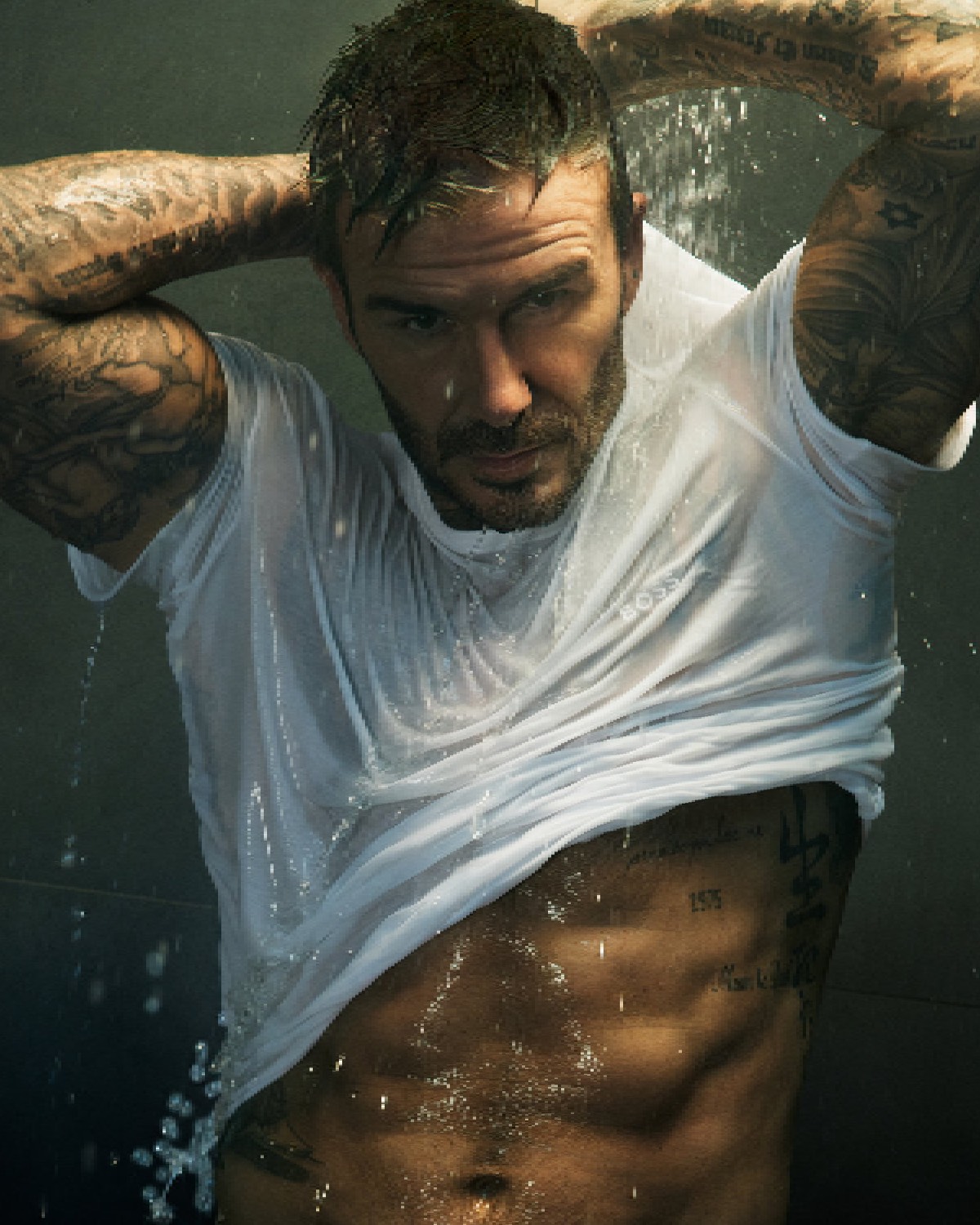 BOSS ONE Bodywear: Mężczyzna w białej, mokrej koszulce BOSS, przez którą prześwituje umięśniona sylwetka. Strumienie wody spływają po jego ciele, dodając sensualnego i dynamicznego efektu. Oświetlenie wydobywa szczegóły tatuaży i fakturę skóry, tworząc dramatyczny, pełen emocji kadr.