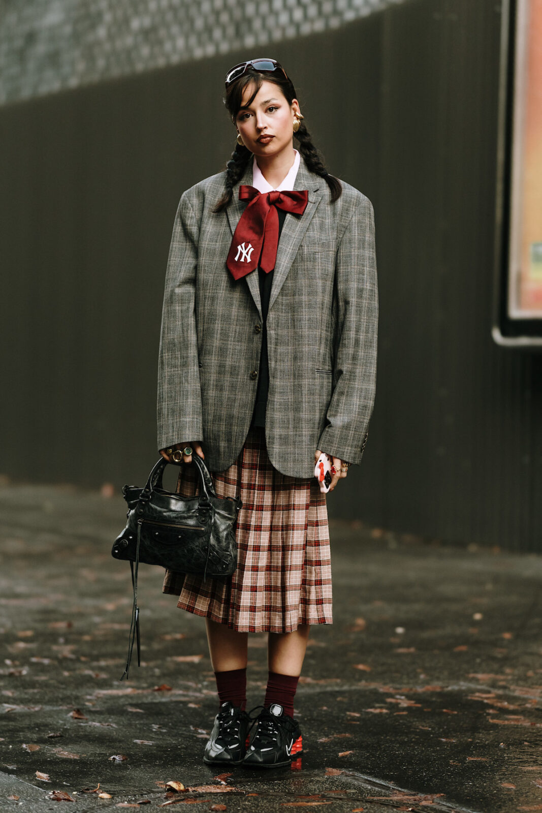 Młoda kobieta w stylowej, eklektycznej stylizacji. Ma na sobie szarą, oversize’ową marynarkę w kratę, bordowy krawat z logo New York Yankees, białą koszulę i plisowaną spódnicę w czerwono-brązową kratę. Na nogach czarne sportowe buty z czerwonymi detalami i bordowe skarpetki. W ręku trzyma czarną skórzaną torebkę z ozdobnymi detalami. Jej włosy są splecione w dwa warkocze, a na głowie ma okulary przeciwsłoneczne. Całość stylizacji uzupełniają złote kolczyki i liczne pierścionki. Tło to miejska przestrzeń z betonowym chodnikiem i metalową ścianą.