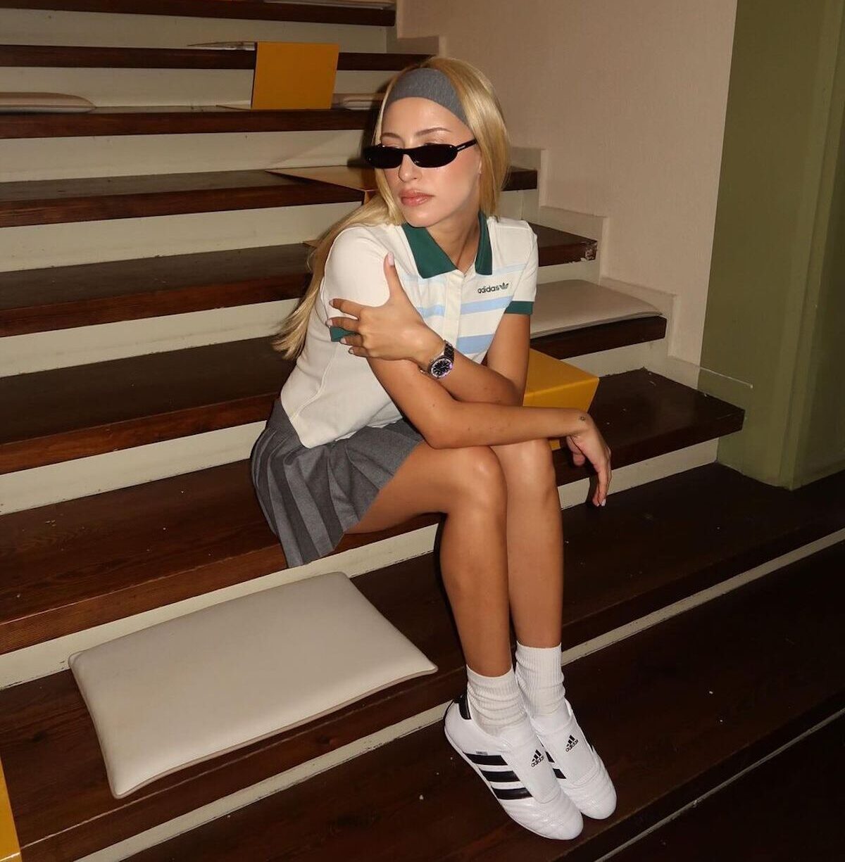 Młoda kobieta stylowo siedzi na drewnianych schodach. Ma na sobie sportowy i modny strój, który składa się z białej koszulki polo Adidas z zielonymi detalami, szarej plisowanej spódniczki oraz białych butów Adidas Taekwondo z czarnymi paskami. Stylizację uzupełnia szara opaska na włosy, wąskie czarne okulary przeciwsłoneczne i srebrny zegarek. W tle widać schody z miękkimi poduszkami i minimalistyczny wystrój wnętrza.