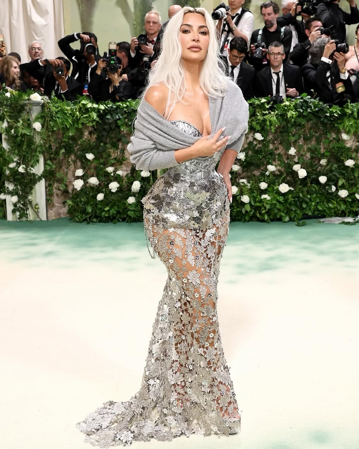 Met Gala Najbardziej kontrowersyjne stylizacje: Kobieta o platynowych włosach ubrana w efektowną, połyskującą suknię z kwiatowymi aplikacjami i transparentnymi wstawkami, owinięta szarym szalem. W tle widoczni fotografowie i ściana zieleni, nadające scenie ekskluzywnego charakteru.