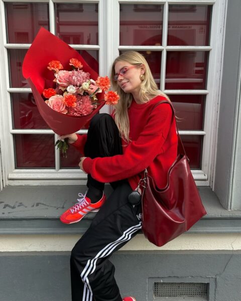 Trendy Pinterest 2025: Kobieta ubrana w czerwony sweter, torebkę i buty, które zestawiła z czarnymi spodniami Adidas, pozująca z bukietem soczystych kwiatów. Siedzi na murku i uśmiecha się.