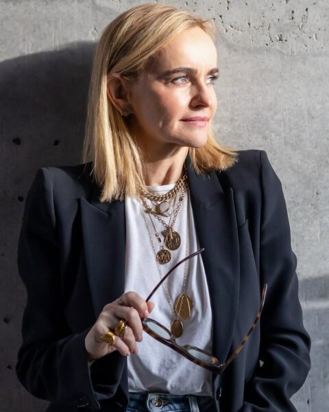 Joanna Czech kim jest: Kobieta w eleganckim czarnym żakiecie i białym topie, ozdobiona złotą biżuterią, patrzy w dal, trzymając okulary w ręce.