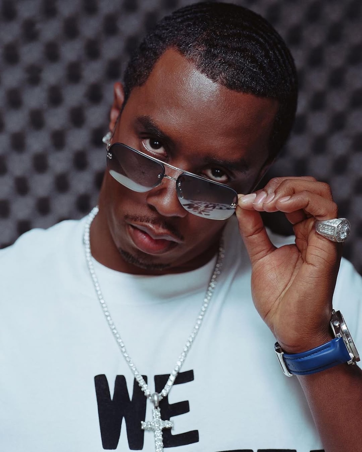 P.Diddy dokument: Raper w białej koszulce z napisem, noszący eleganckie okulary przeciwsłoneczne i biżuterię, patrzy spod okularów z poważnym wyrazem twarzy.