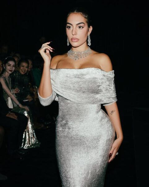 Georgina Rodriguez WAG: Kobieta w srebrnej sukni z odkrytymi ramionami, ozdobiona błyszczącą biżuterią, pozuje na ciemnym tle.