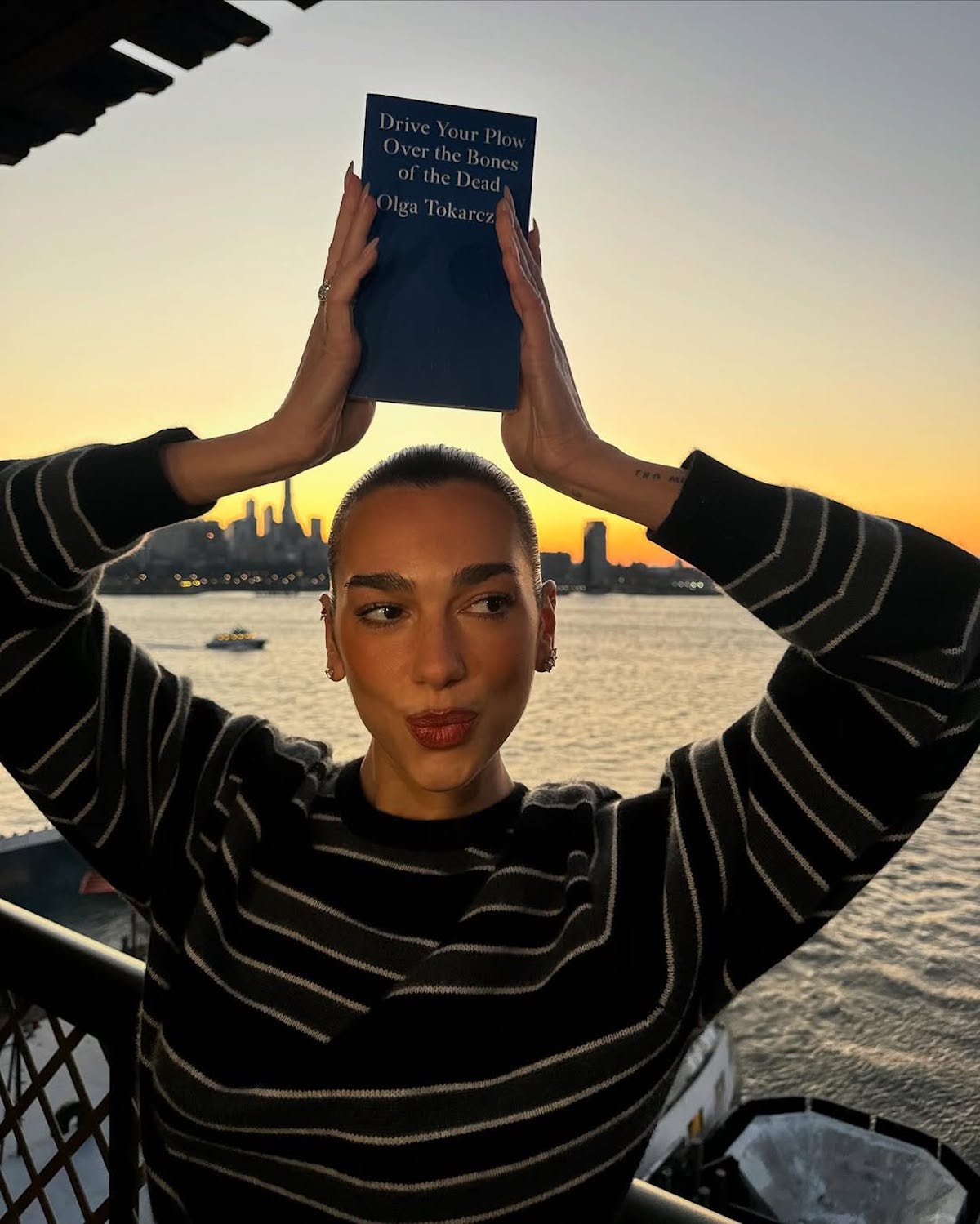 Dua Lipa w czarno-białym swetrze w paski trzyma książkę nad głową, na tle zachodzącego słońca nad rzeką. Okładka książki jest niebieska i zawiera tytuł 'Drive Your Plow Over the Bones of the Dead' autorstwa Olgi Tokarczuk. W tle widać łódź na wodzie oraz zarys miejskiego krajobrazu.