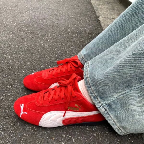Najmodniejsze sneakersy 2025 – czerwone Puma Speedcat z białymi detalami i logotypem, widoczne na tle asfaltu. Buty mają sportowy design z akcentami w stylu retro, idealnie komponując się z szerokimi, jasnoniebieskimi jeansami.