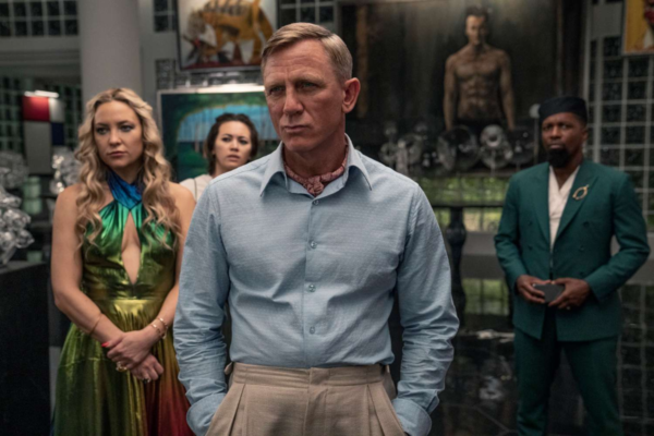 Premiery Netflix 2025: Cztery osoby stojące w eleganckim, nowoczesnym pomieszczeniu w galerii sztuki. Na pierwszym planie mężczyzna w jasnoniebieskiej koszuli z apaszką i beżowych spodniach, wyraziste spojrzenie skierowane w bok. W tle po lewej stronie kobieta w kolorowej sukience z głębokim dekoltem, a obok niej inna kobieta w białym stroju z zaniepokojoną miną. Po prawej stronie mężczyzna w zielonym garniturze i beretce. W tle widoczne dzieła sztuki oraz rzeźby.