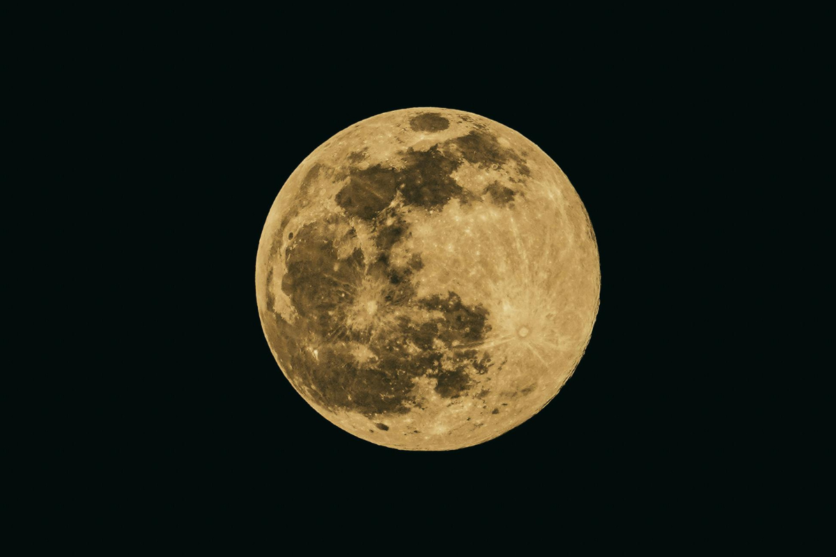 Zdjęcie przedstawia pełnię Księżyca na ciemnym, nocnym niebie. Księżyc jest w pełni oświetlony, widoczne są wyraźne szczegóły jego powierzchni, takie jak kratery i ciemniejsze obszary morz księżycowych. Całość utrzymana w złocistych odcieniach.