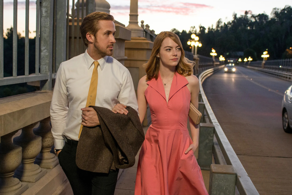 „Scena z filmu „La La Land” przedstawiająca Ryana Goslinga i Emmę Stone spacerujących po oświetlonym moście o zmierzchu. Gosling ubrany jest w białą koszulę, żółty krawat i trzyma brązową marynarkę, podczas gdy Stone ma na sobie różową sukienkę z rozkloszowanym dołem i trzyma kopertówkę. W tle widać latarnie, mostowe balustrady i nadjeżdżające samochody.”