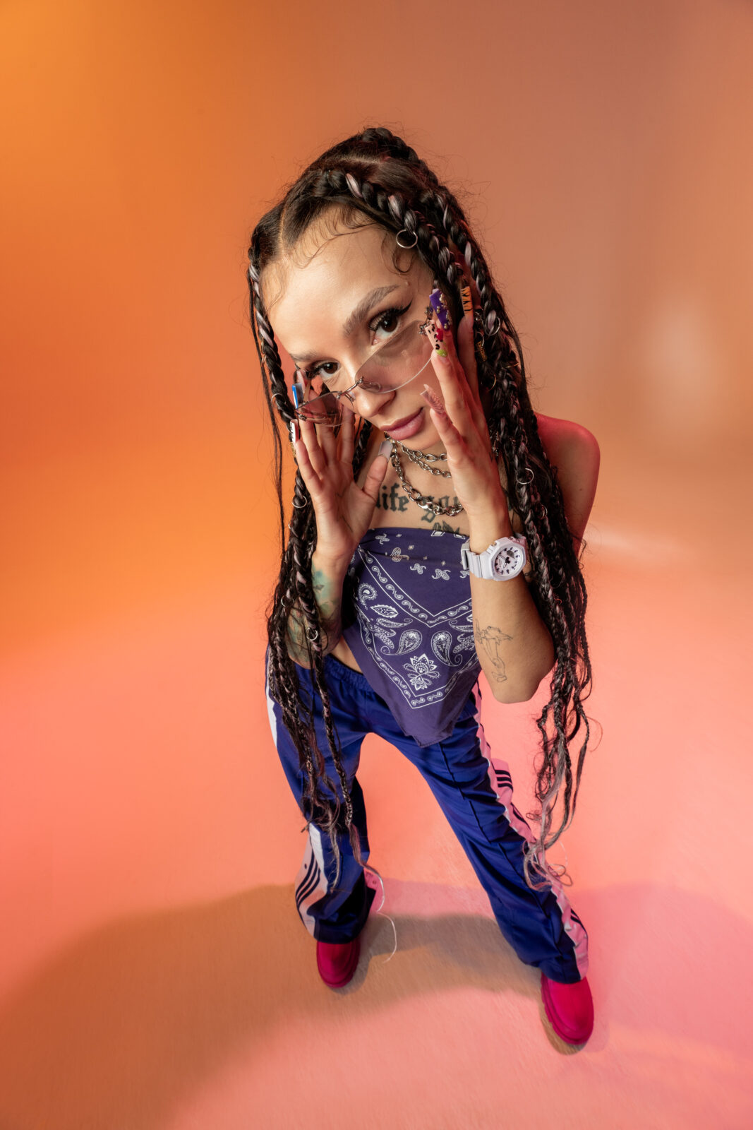 Modelka w fioletowej bandanie i sportowych spodniach Adidas z odważnymi warkoczami ozdobionymi pierścieniami. Jej paznokcie zdobione są kolorowymi wzorami, a dodatki – łańcuchy, zegarek G-Shock oraz okulary – podkreślają jej indywidualny styl. Pomarańczowe tło kontrastuje z chłodnymi tonami jej ubioru.