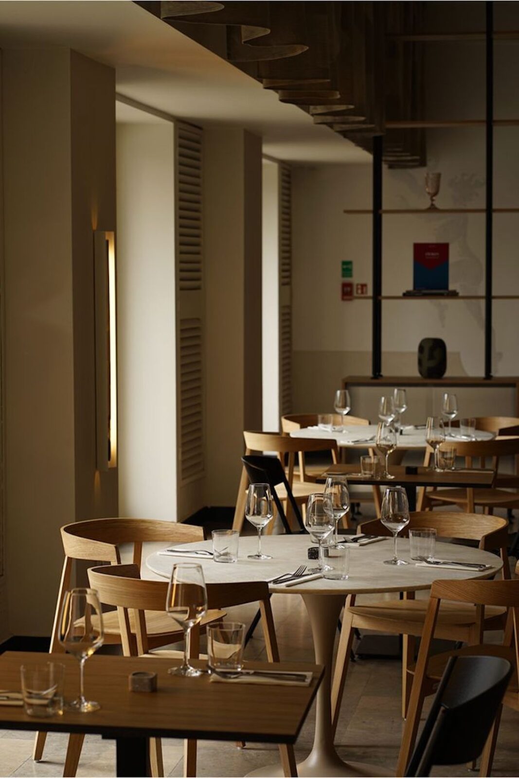 Stylowe wnętrze nowoczesnej restauracji z eleganckimi, minimalistycznymi drewnianymi krzesłami i okrągłymi oraz prostokątnymi stołami nakrytymi jasnymi obrusami. Na stołach starannie ułożone kieliszki do wina, szklanki oraz sztućce, co nadaje miejscu wyrafinowany charakter. Ciepłe, subtelne oświetlenie oraz naturalne kolory drewna i beżu tworzą przytulną, kameralną atmosferę. Aranżacja wpisuje się w trendy "nowe restauracje w Warszawie na wiosnę 2025", oferując eleganckie miejsce na wyjątkowe spotkania i kolacje.