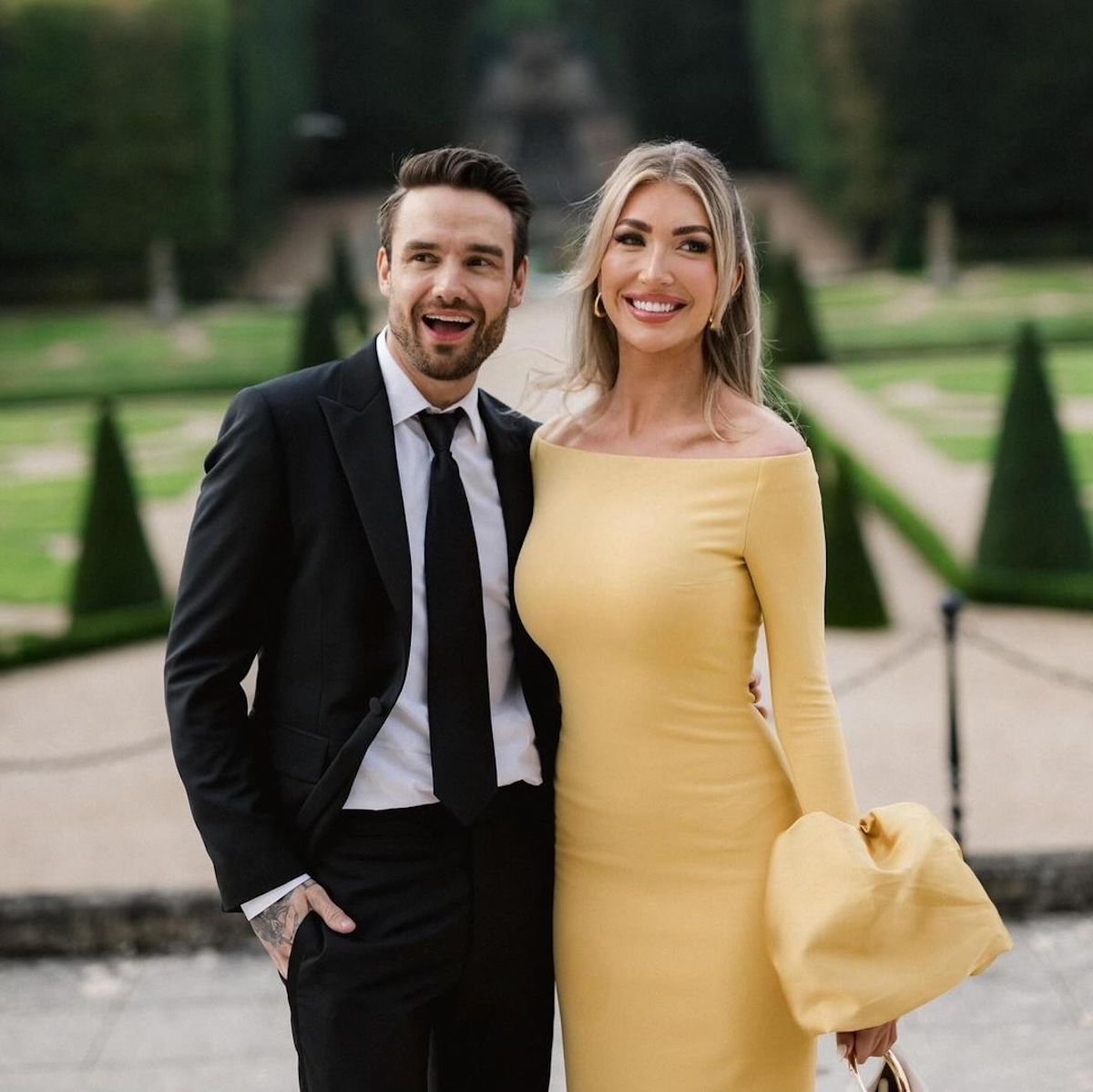 Elegancka para pozująca w ogrodzie formalnym. Mężczyzna w czarnym garniturze z białą koszulą i krawatem uśmiecha się szeroko. Kobieta w żółtej sukni z długimi rękawami również uśmiecha się radośnie. W tle widoczne geometryczne kształty krzewów oraz alejki ogrodowe.