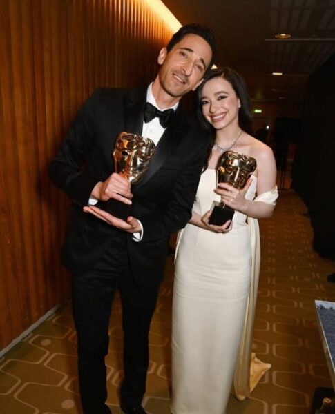 Zwycięzcy BAFTA 2025 z uśmiechem pozują z nagrodami. Mężczyzna w eleganckim czarnym smokingu i kobieta w kremowej, dopasowanej sukni z odkrytymi ramionami prezentują swoje statuetki. Stylizacje BAFTA 2025 zachwycają klasyką i szykiem – on w klasycznej muchy i marynarce, ona w minimalistycznej kreacji z subtelną biżuterią. Tło stanowi elegancka, drewniana ściana, a w ich wyrazie twarzy widać radość i dumę.