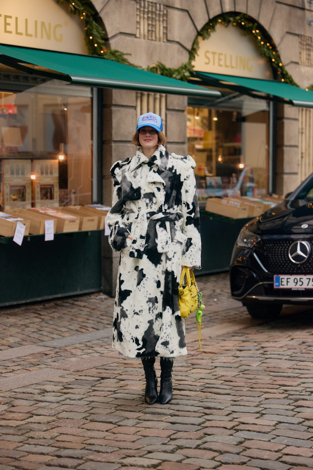 Kobieta pozuje na brukowanej ulicy w długim płaszczu z futrzanym wykończeniem w biało-czarny wzór przypominający krowie łatki, czyli cow print na wiosnę 2025. Płaszcz jest przewiązany w talii paskiem i ma efektowne bufiaste ramiona. Stylizację uzupełniają czarne botki na obcasie, jasnoniebieska czapka z daszkiem oraz żółta torebka z brelokiem w kształcie zielonej żabki.