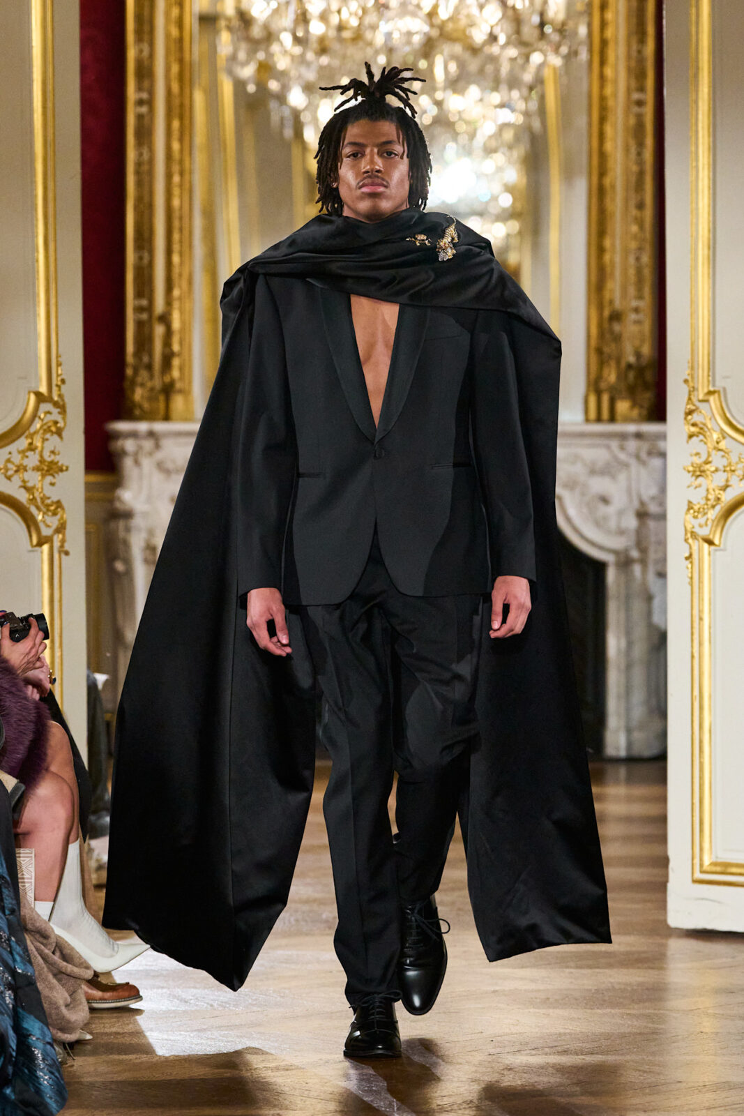 Henry Samuel kto to: Model w czarnym garniturze z peleryną ozdobioną broszką – Model prezentuje niezwykle elegancki, czarny garnitur z dramatycznym, długim płaszczem narzuconym na ramiona, ozdobionym błyszczącą broszką w kształcie ptaka. Jego poważne spojrzenie i głęboki dekolt marynarki podkreślają teatralny charakter stylizacji.
